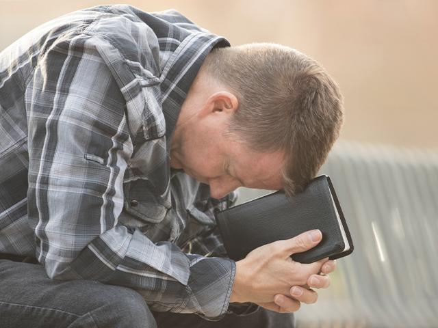 man bending over on a bench with Bible in hands praying