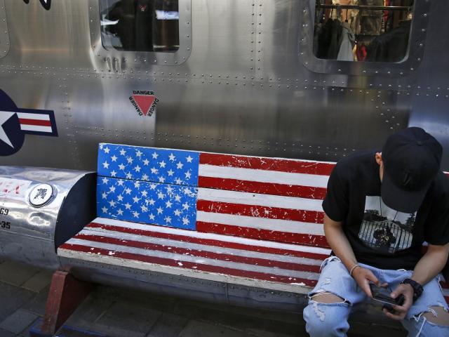 A man browses his smartphone on a bench with a decorated with U.S. flag outside a fashion boutique selling U.S. brand clothing at the capital city&#039;s popular shopping mall in Beijing, Monday, May 13, 2019. (AP Photo/Andy Wong)