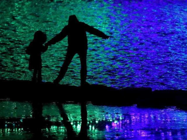 In this Nov. 30, 2018, file photo people are silhouetted against holiday lights reflecting off a pond in a park in Lenexa, Kan. (AP Photo/Charlie Riedel, File) 