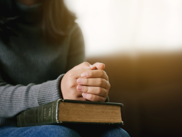 person with her hands clasped atop a closed Bible in her lap 