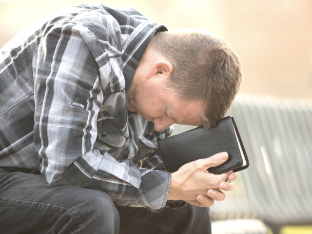 man in a plaid shirt bowing his head on his Bible
