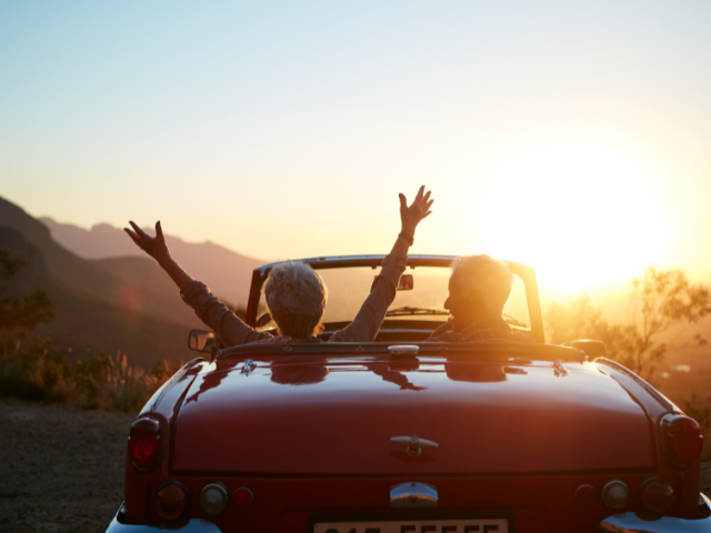 woman in a convertible raising arms with praise as sun sets in the mountains