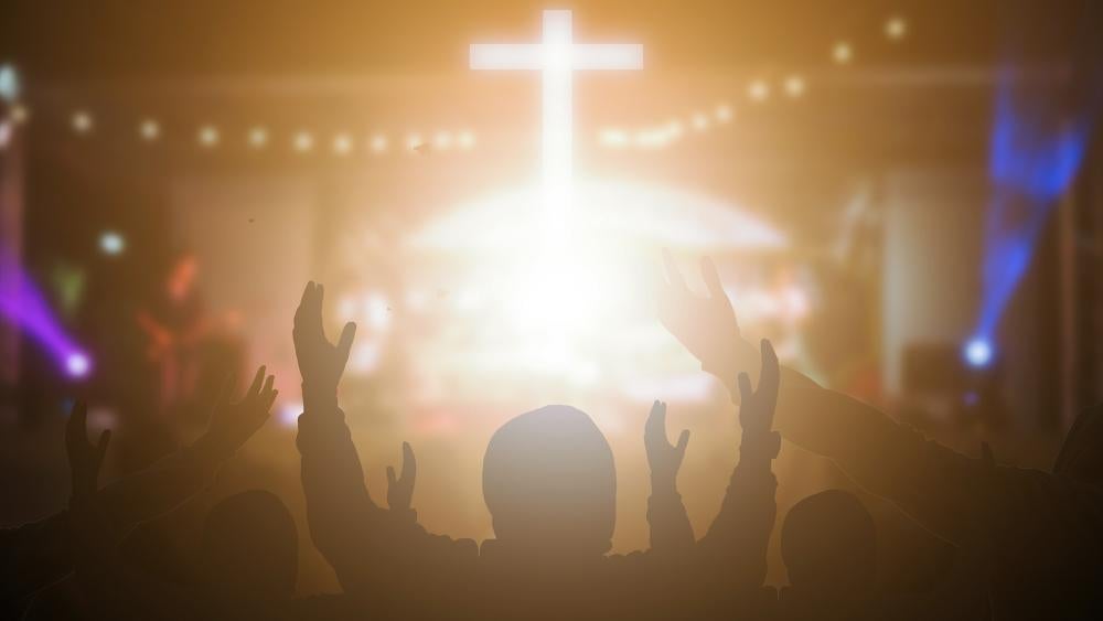 Christian churchgoers raise their hands in worship to Jesus as a cross shines on stage. (Adobe stock image)